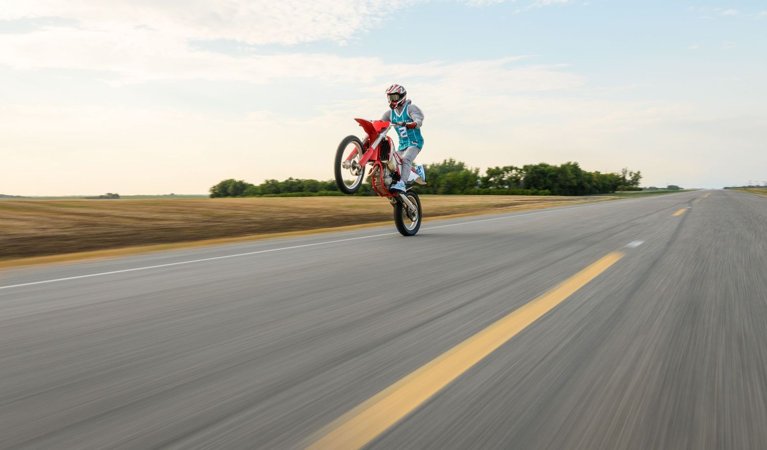 Motorcycle rider performing a wheelie on a red dirt bike on an empty rural highway.
