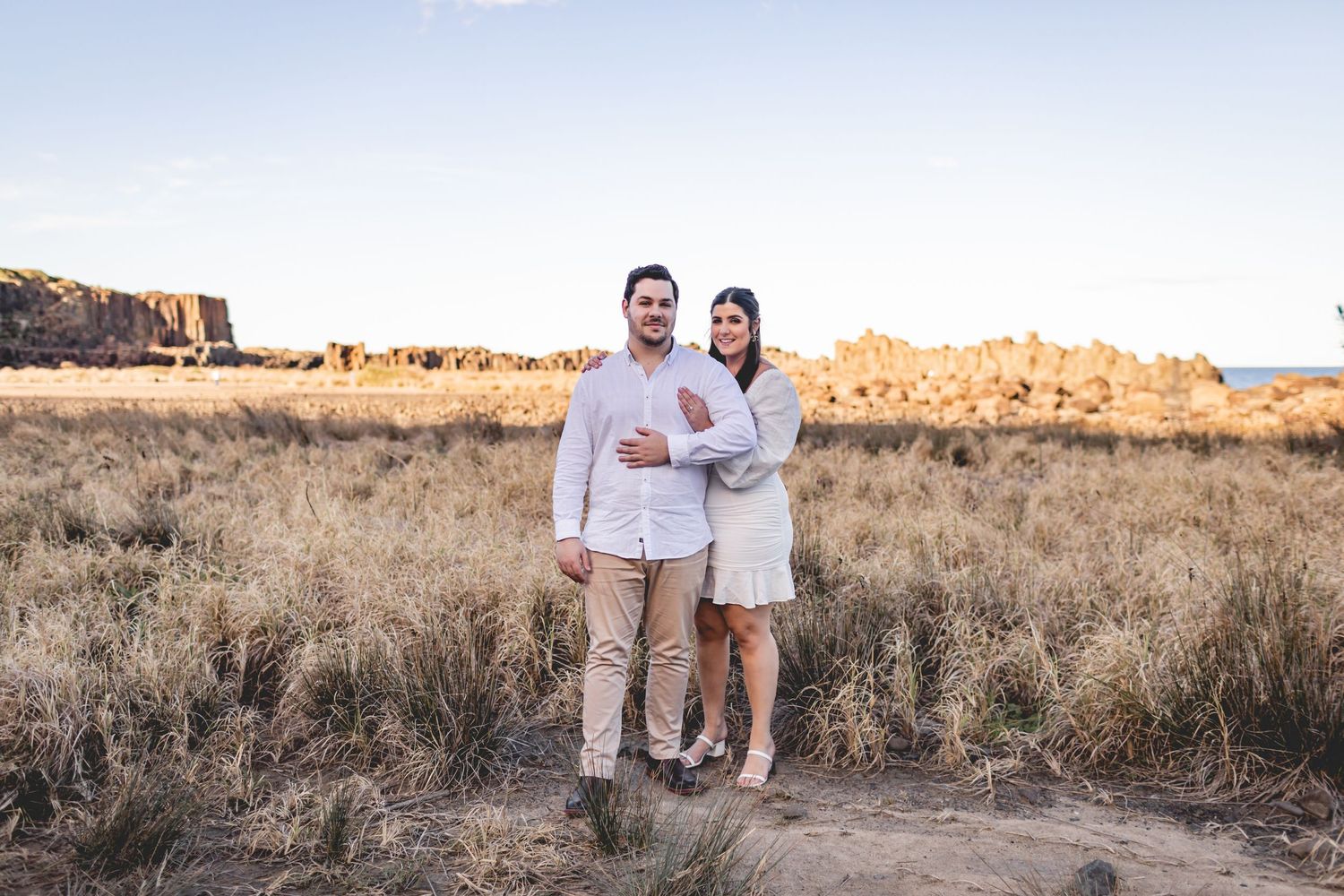 A couple poses together in white outfits against a desert landscape with red cliffs.