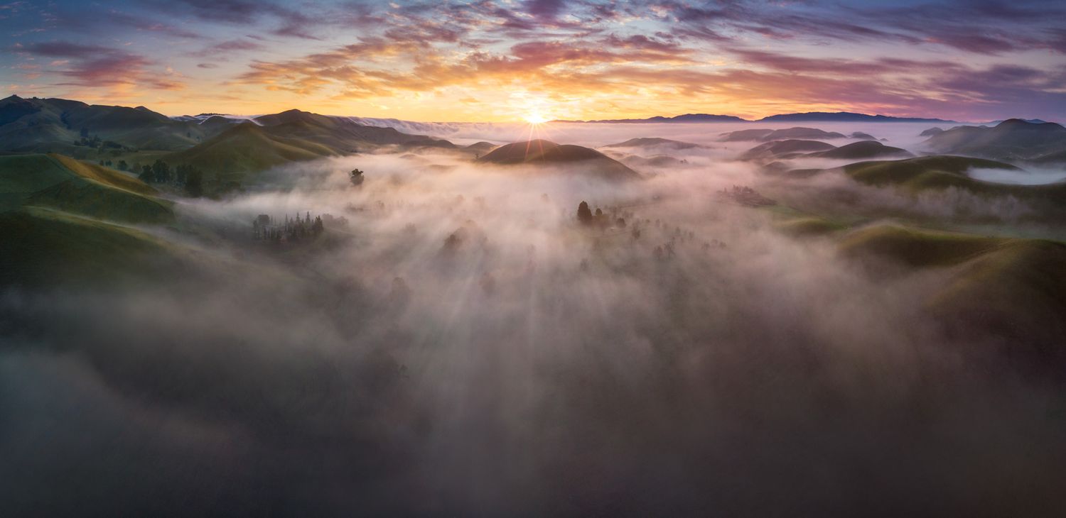Misty mountain valley landscape at sunset with dramatic orange sky and rolling fog between hills.