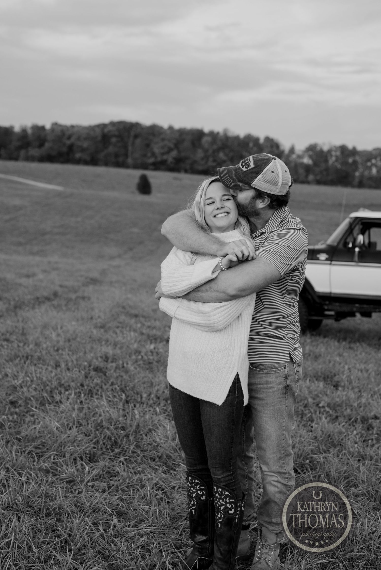 Black and white photo of a couple embracing in a field with a truck in the background.