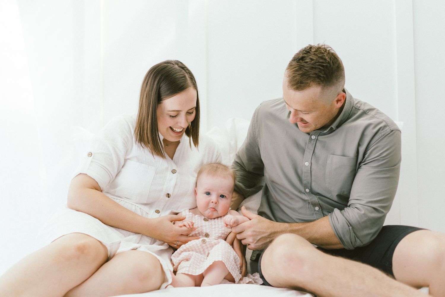 Parents and infant share loving moments together on white bedding during intimate lifestyle photo session.