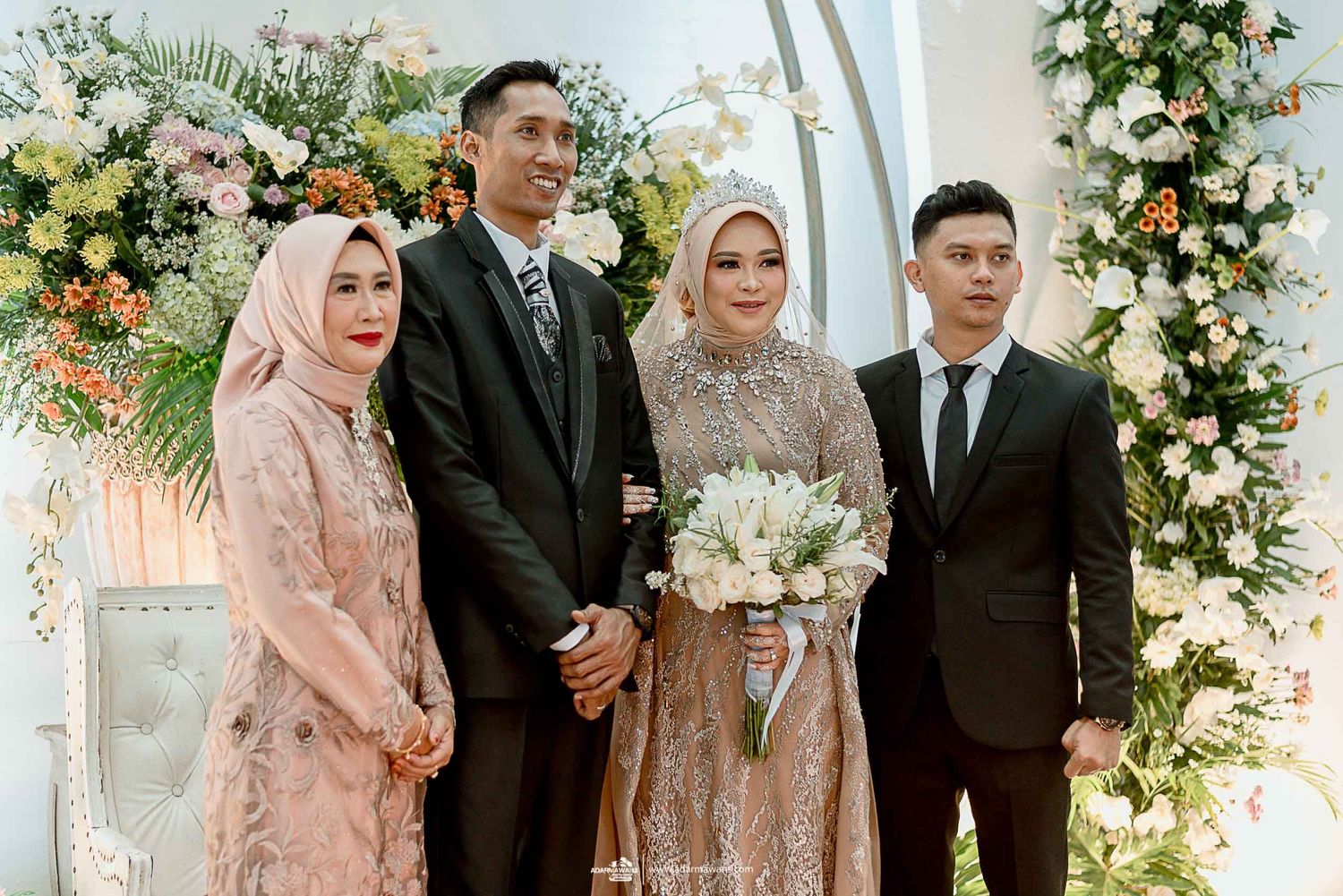 Wedding couple and guests pose under an elegant floral arch with peach and white flower decorations.
