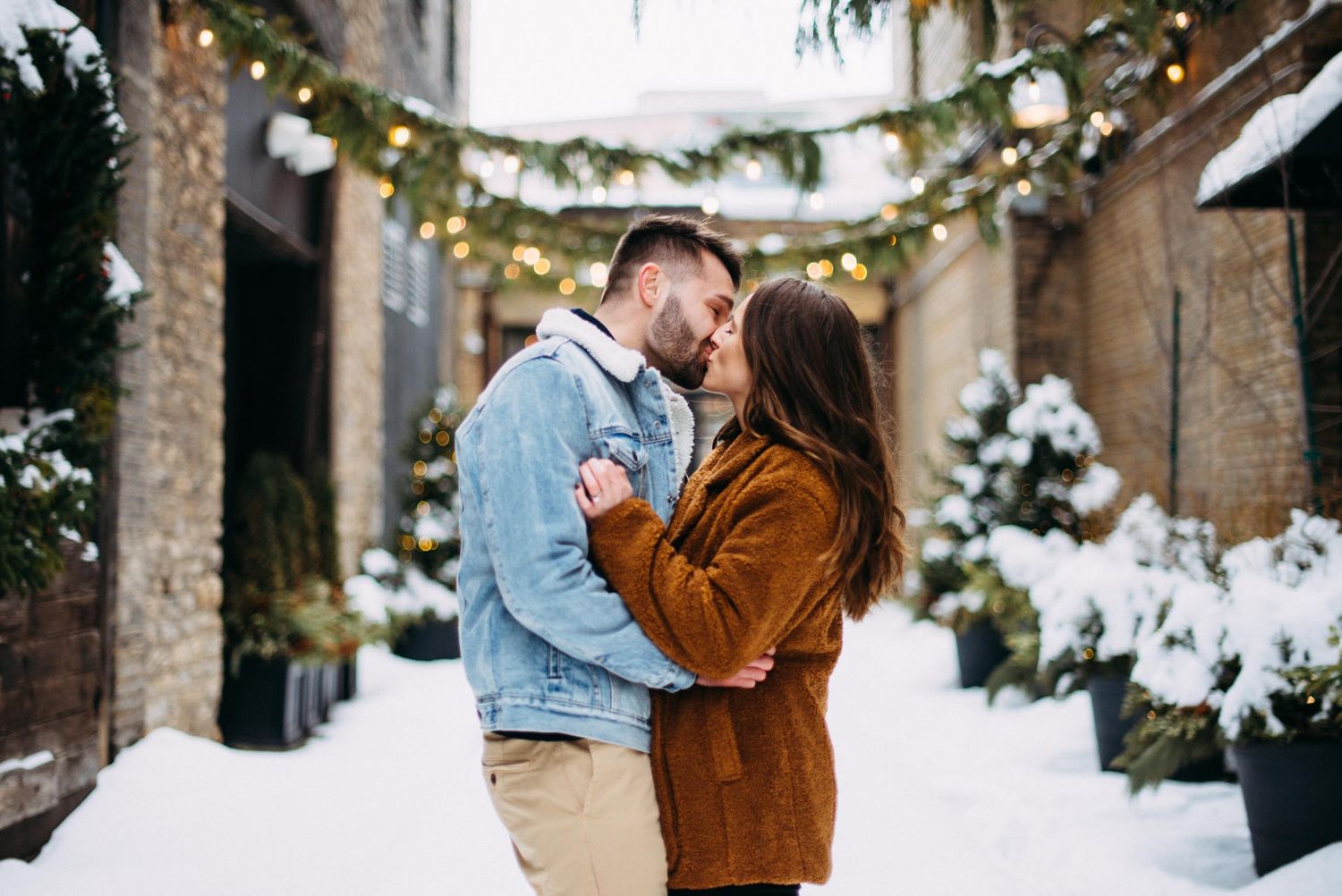 Couple kisses in the snow under holiday lights for their engagement portraits near Hewing Hotel.