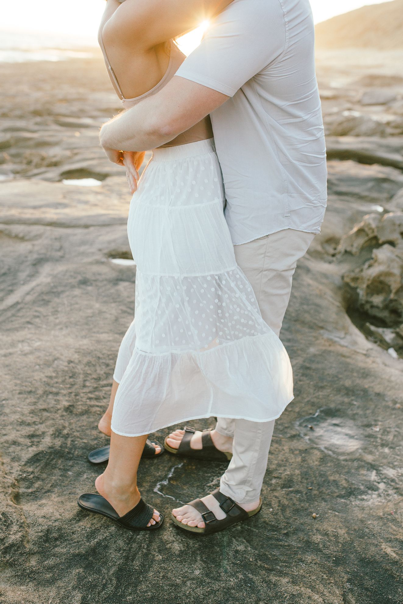 Close up of a couple embracing, showing white dress and sandals details on rocky ground.