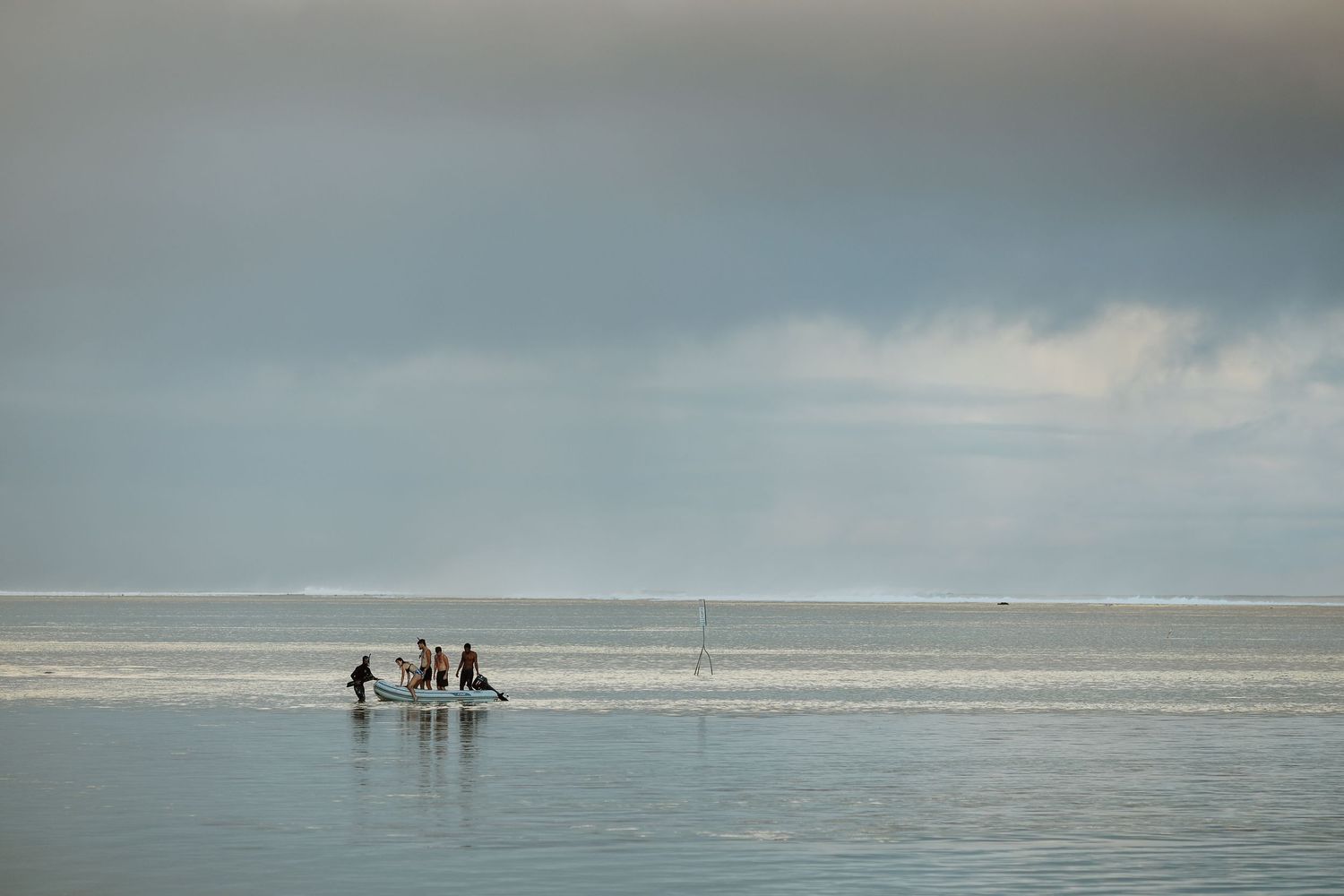 Distant silhouettes stand in shallow ocean water under a moody cloudy sky.
