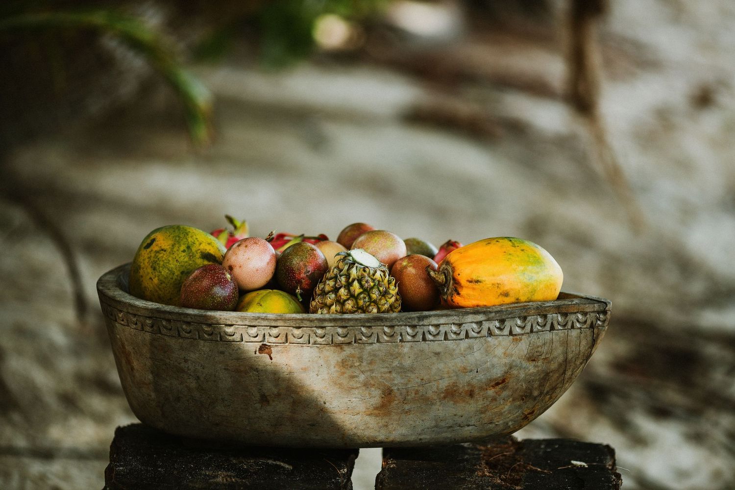 A rustic metal bowl filled with tropical fruits sits in dappled sunlight.