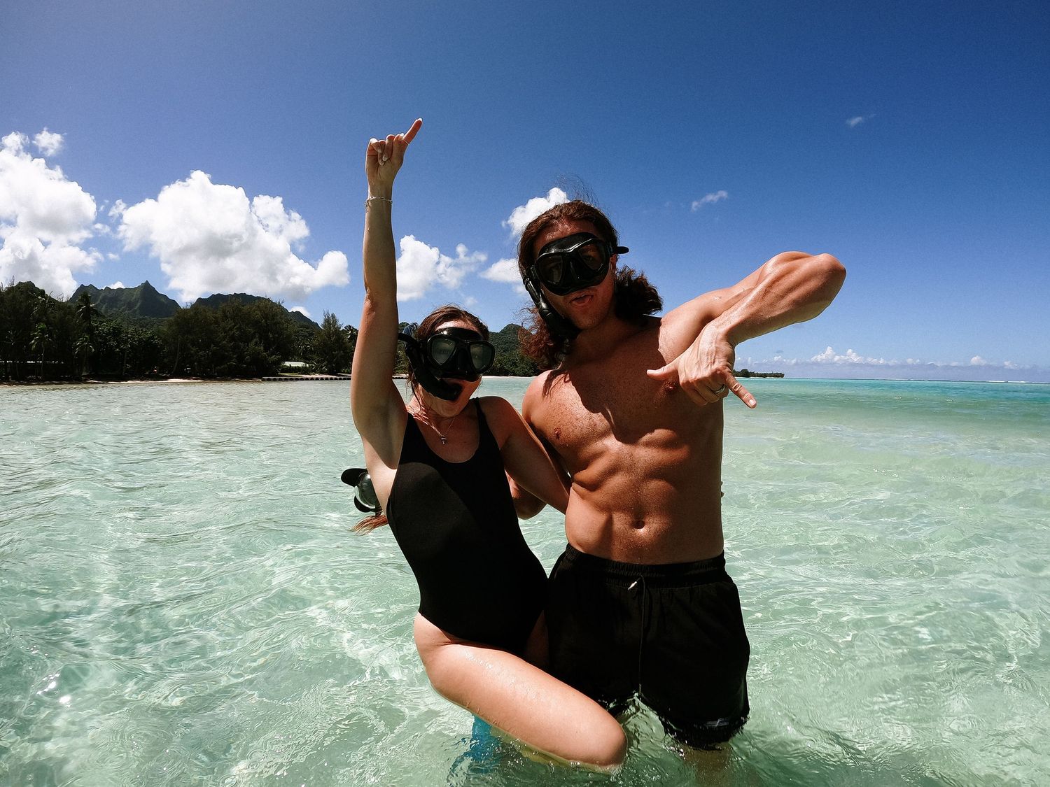 Two people jumping in shallow tropical waters with clear turquoise ocean and beach background.