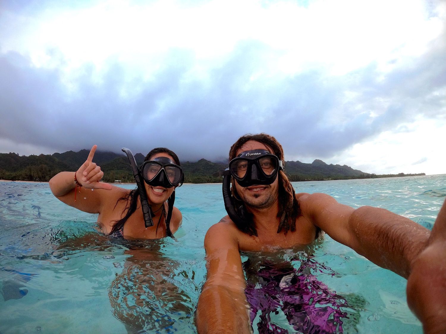 Snorkelers taking selfie in crystal clear ocean waters with cloudy sky overhead.