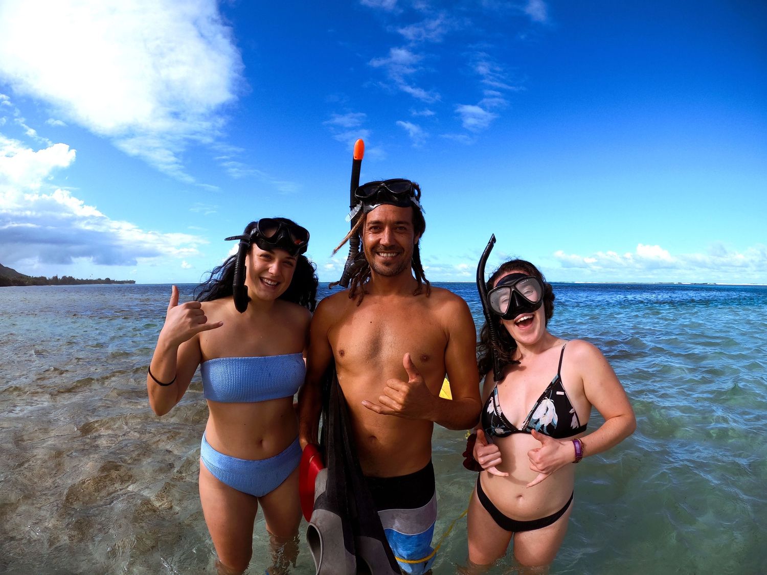 Three snorkelers pose together on tropical beach with blue sky and ocean in background.