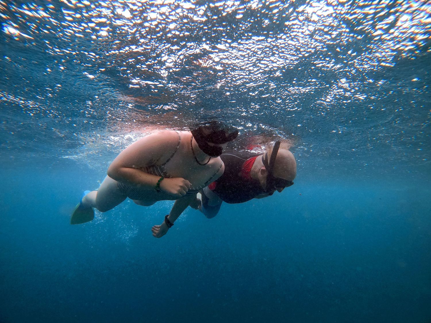 Two snorkelers swim underwater in clear blue tropical waters with sunlight filtering through from above.