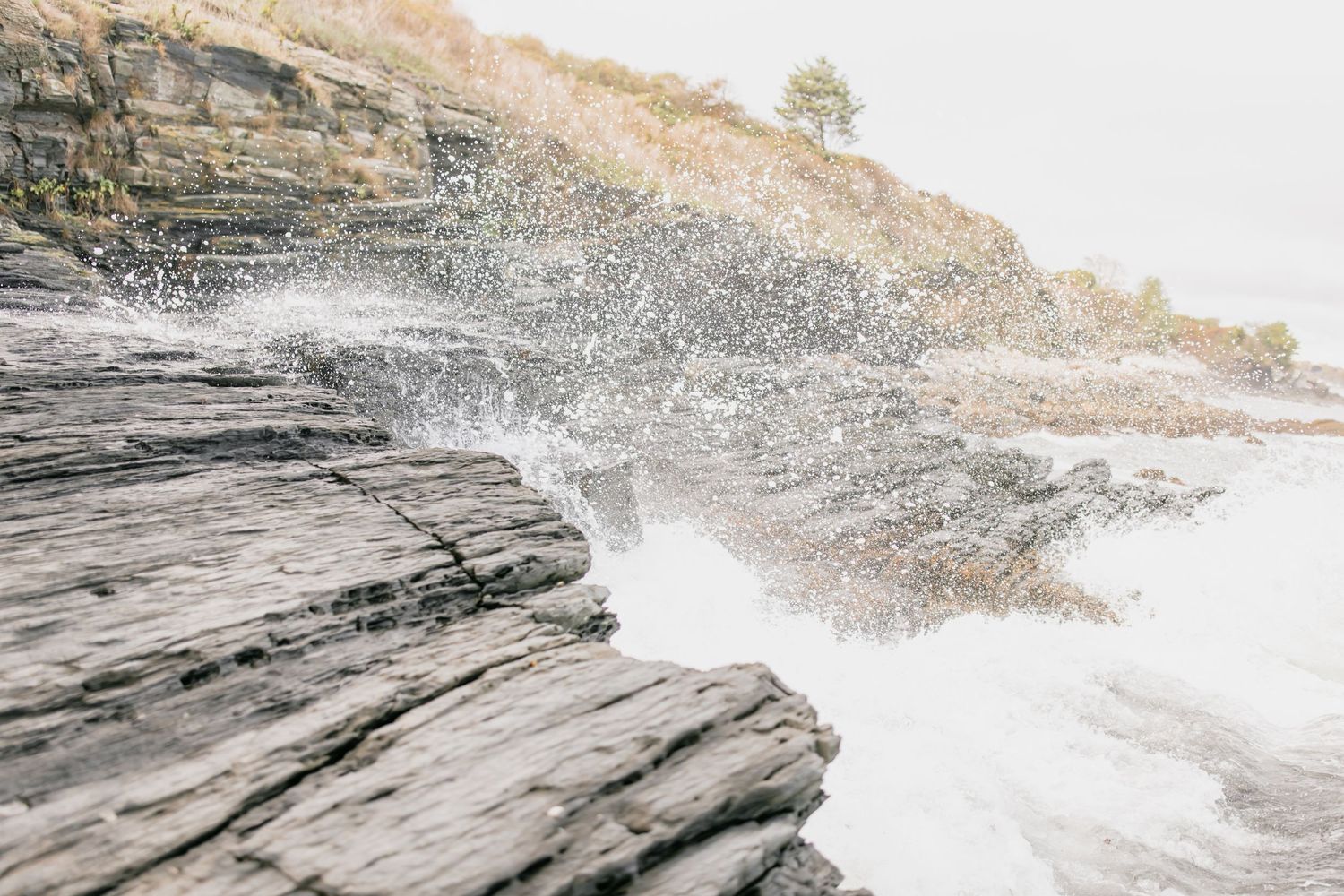 Powerful ocean waves crashing against layered rocky coastal cliffs with sea spray and lush greenery above.