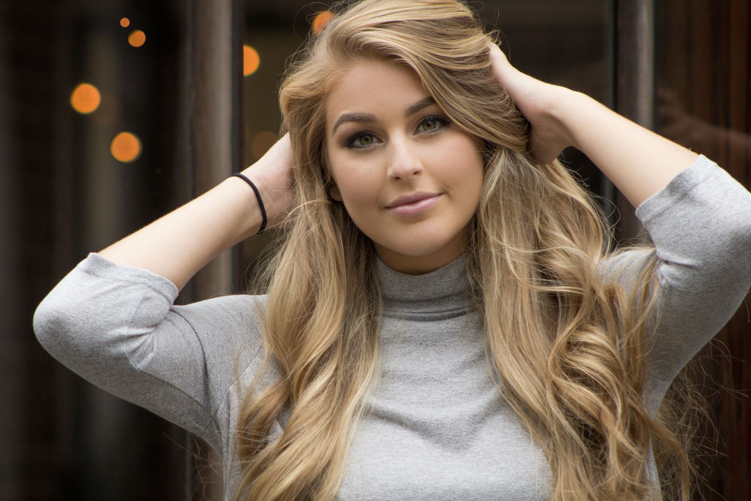 Young blonde woman with long wavy hair wearing a gray turtleneck, posing with hands behind her head.