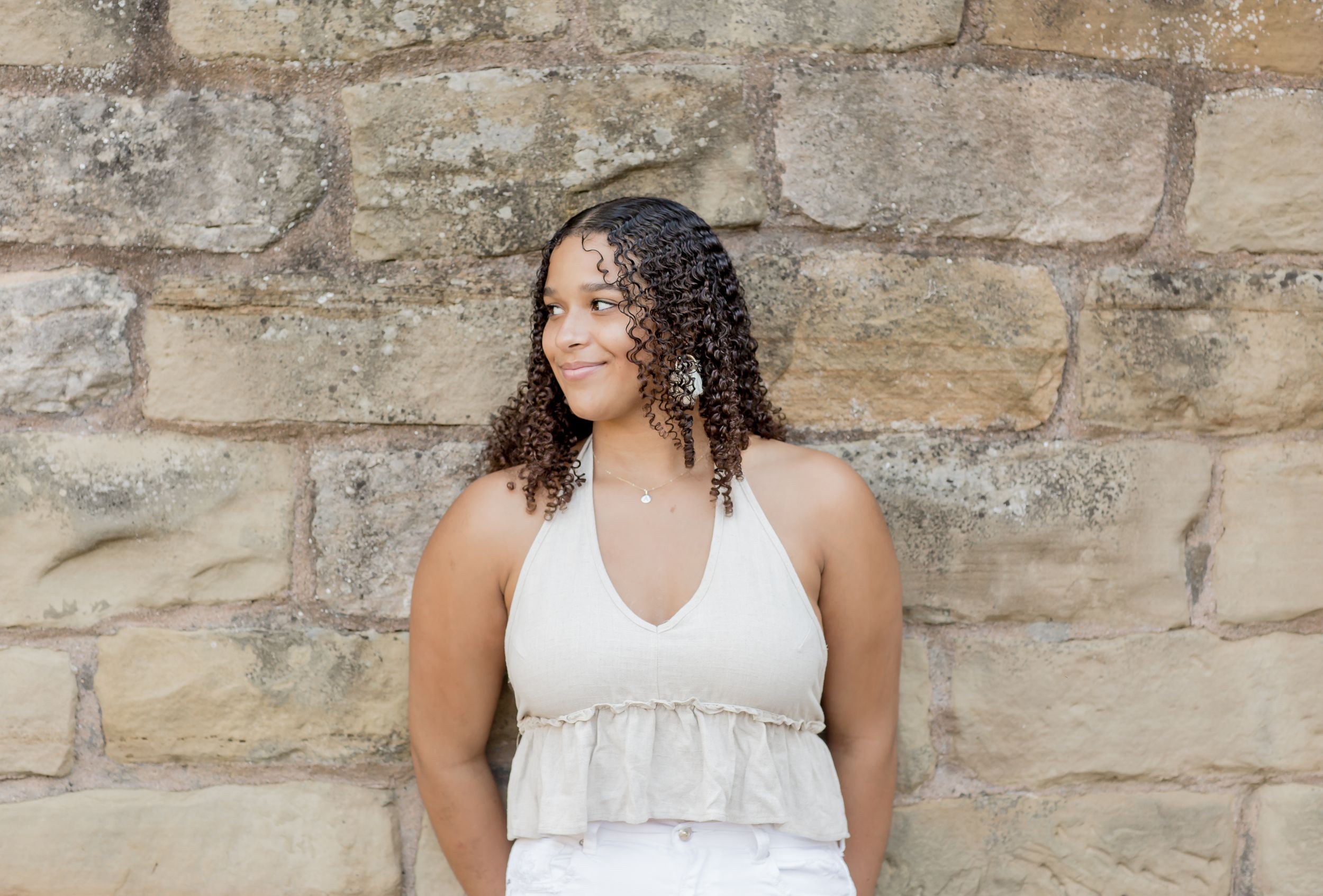 Young woman with curly hair in a white halter top smiling outdoors against a rustic stone wall.