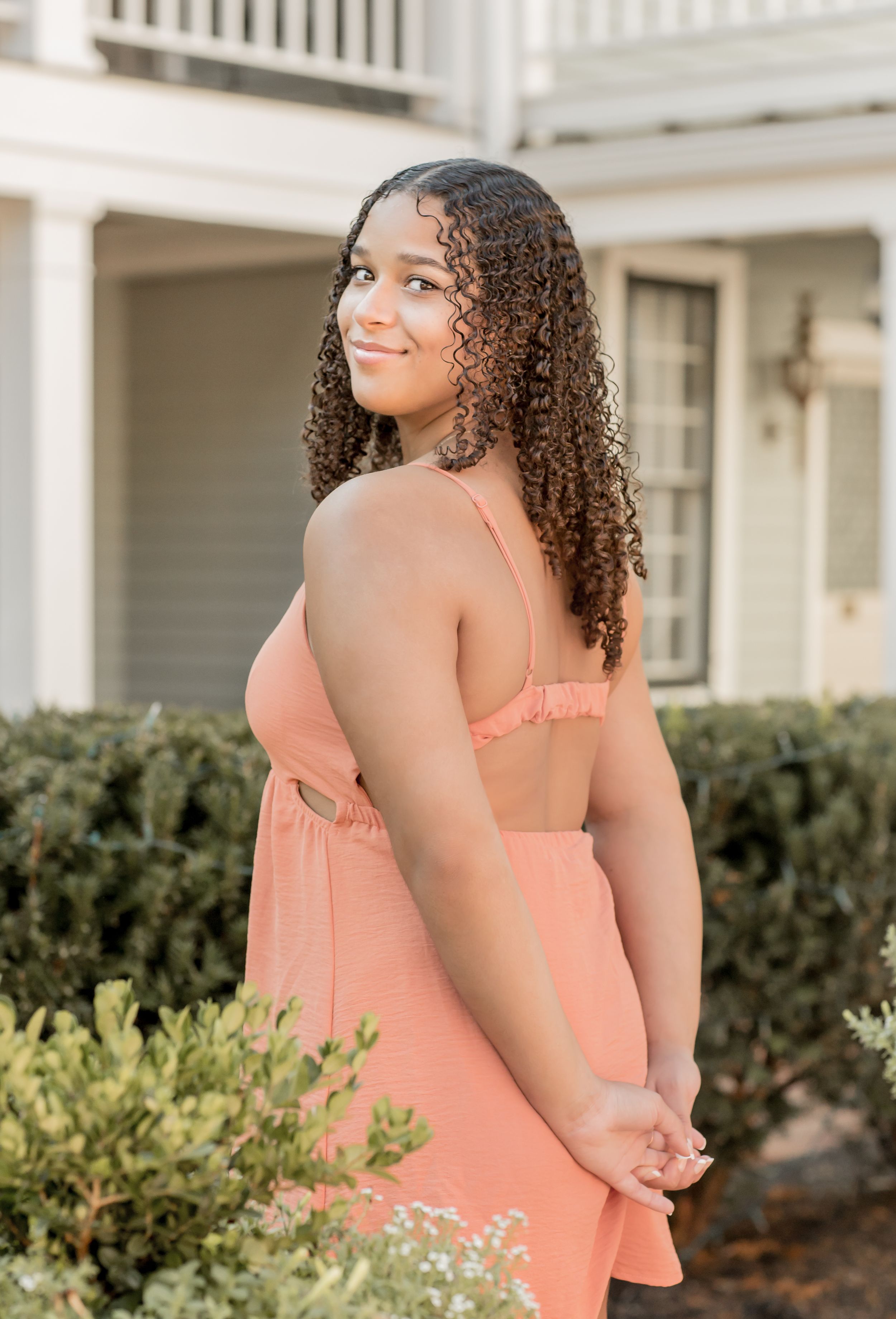 Young woman in a coral dress with curly hair smiling over her shoulder outdoors near a house.