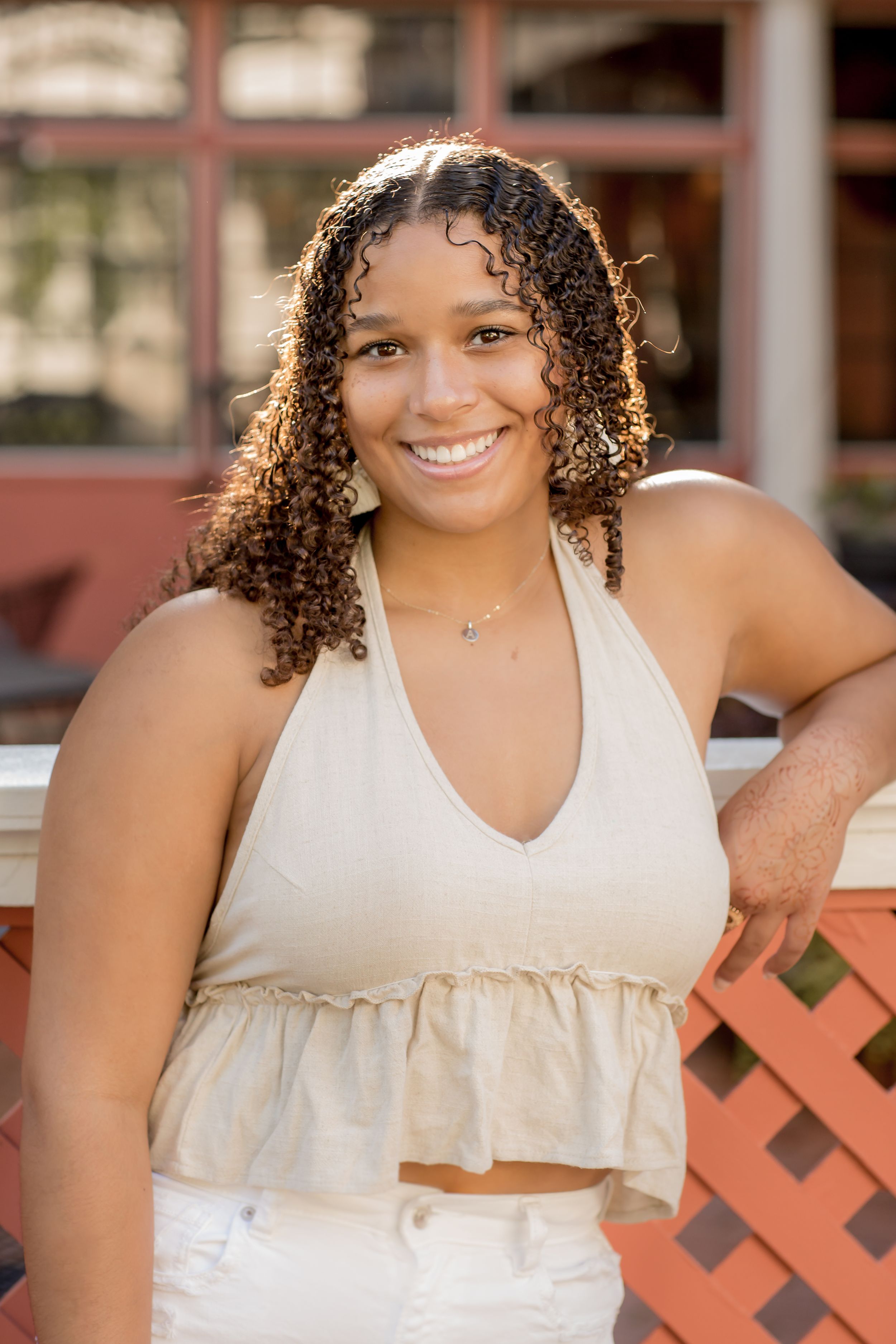 Smiling young woman with curly hair wearing a beige halter crop top and white jeans, posing outdoors.