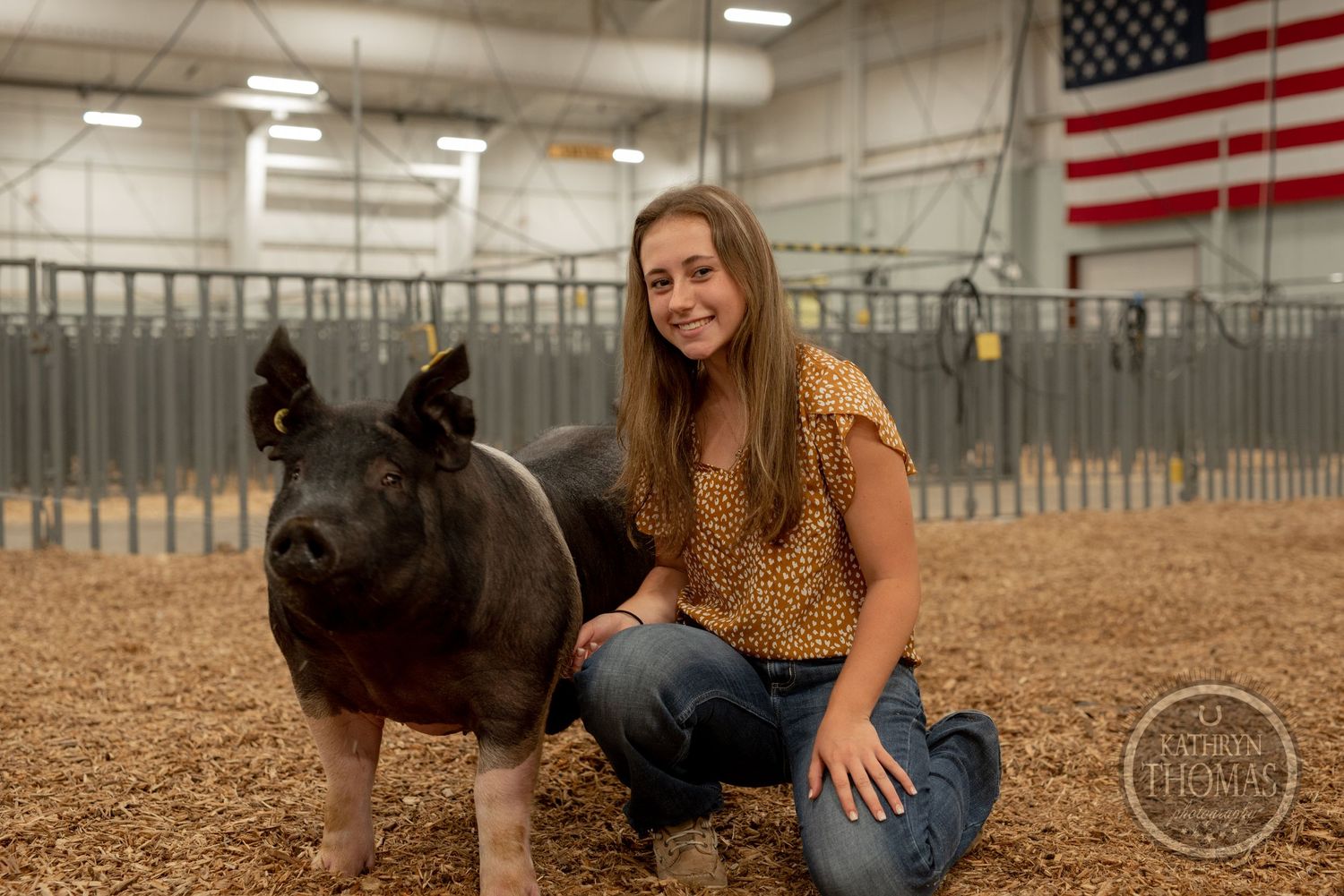 Black show pig poses with handler in sawdust-covered pen with American flag displayed.