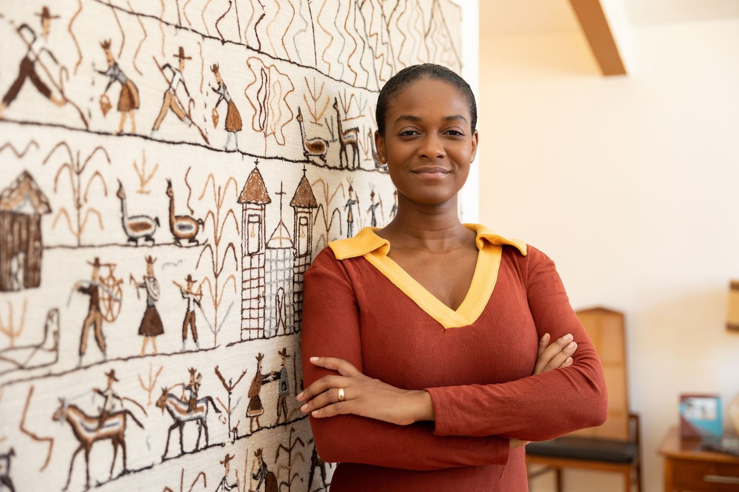 A confident Black woman in a red and gold top stands with arms crossed before an African tapestry wall art.