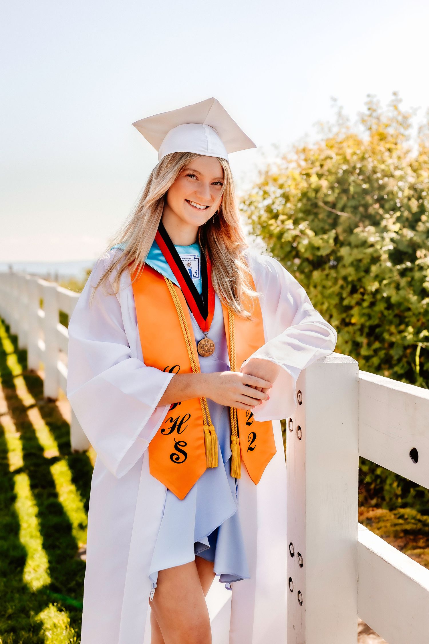 Graduate wearing white cap and orange stole poses by white fence on sunny day.