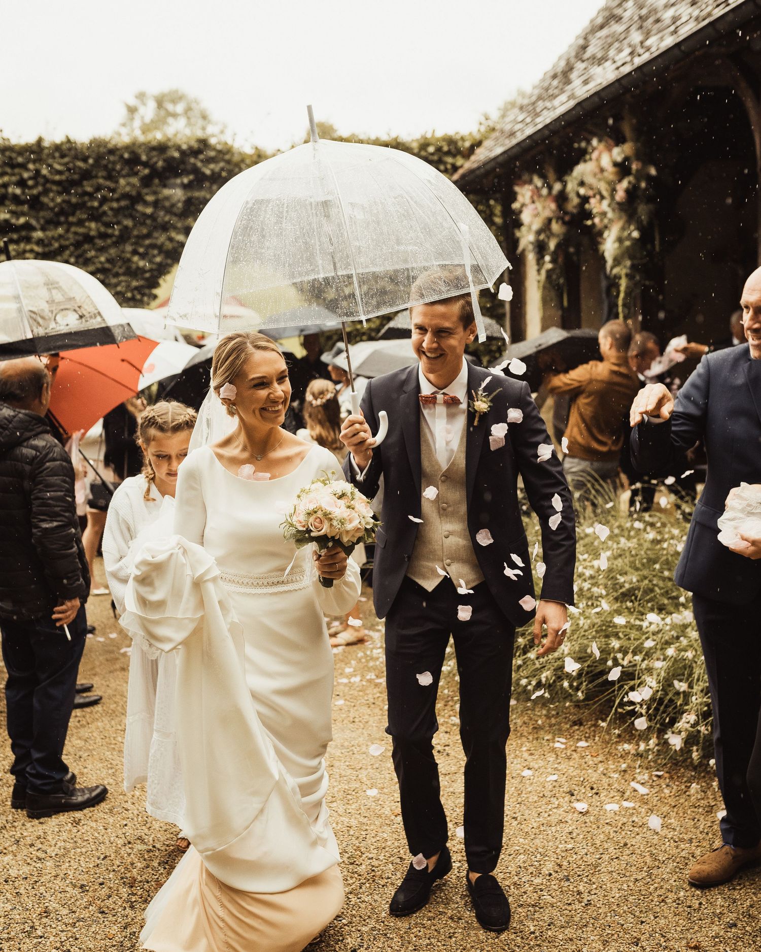 Wedding party walking under clear umbrellas on rainy garden path.