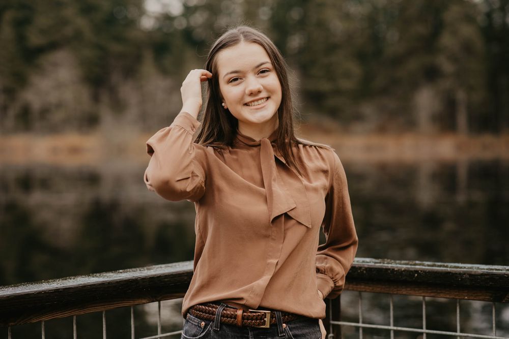 Woman in tan blouse smiling while standing on bridge with lake backdrop.