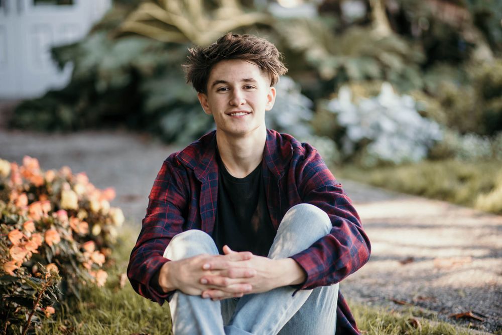 Young person in plaid shirt sitting casually outdoors near flowering bushes.