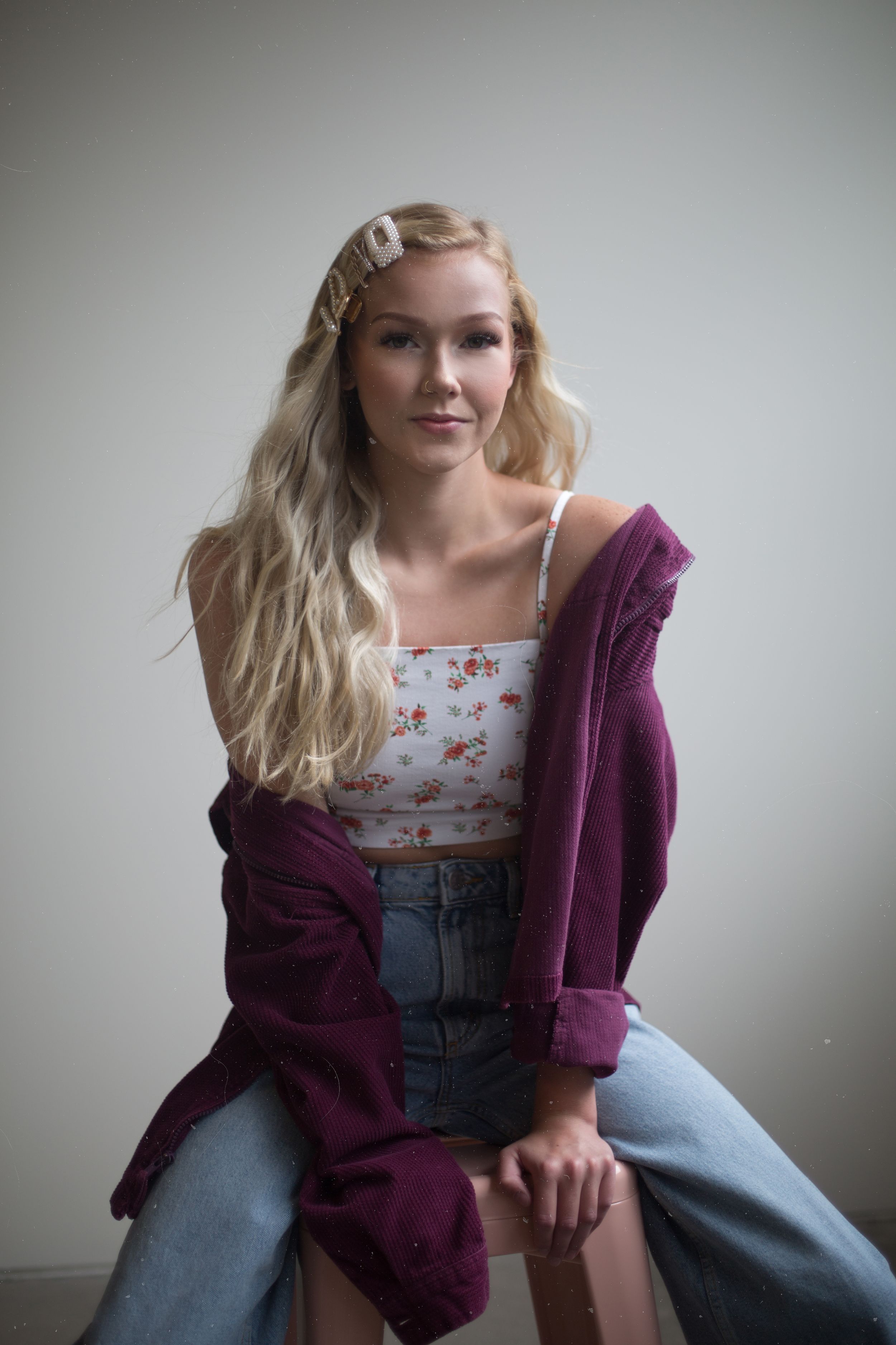 Person in purple cardigan and floral top poses on chair against light background.