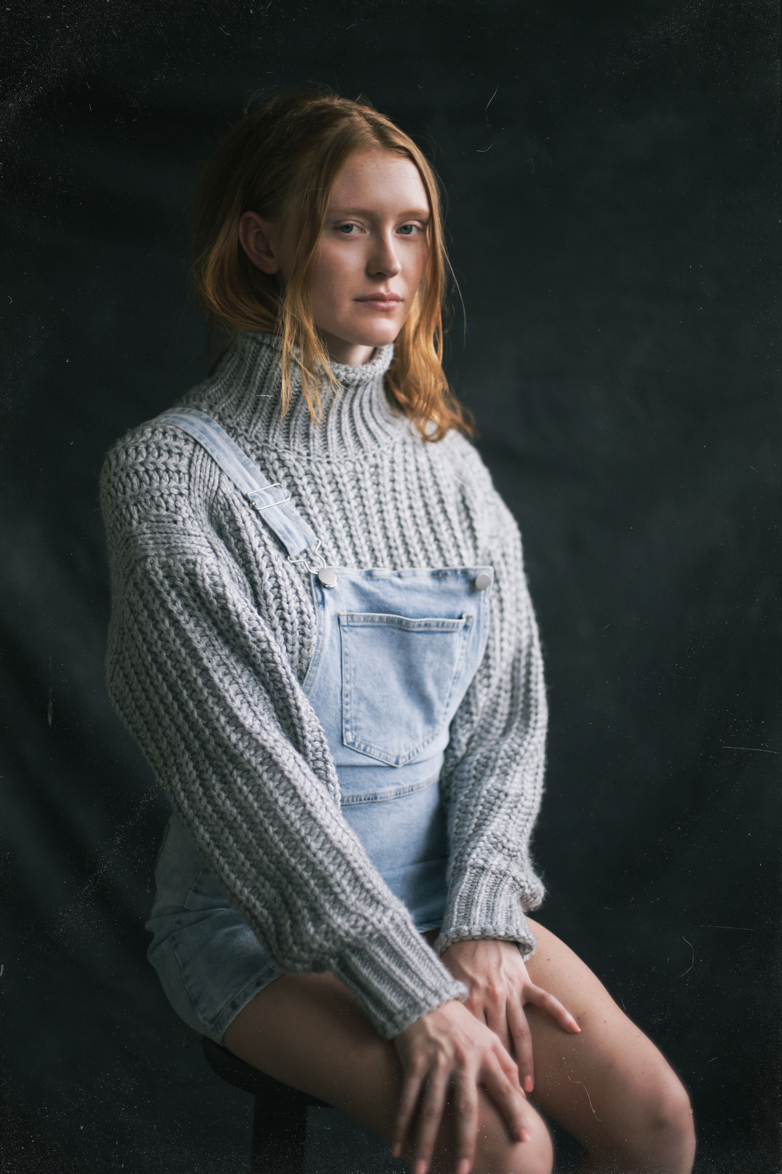 Model in cozy gray knit sweater and denim overalls poses against dark background in artistic portrait series.