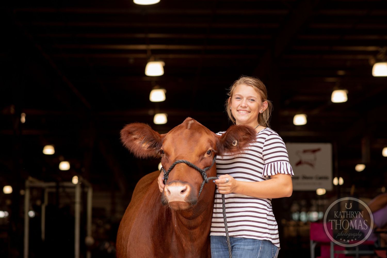 A person in a striped shirt stands with their brown show cow under indoor lighting.