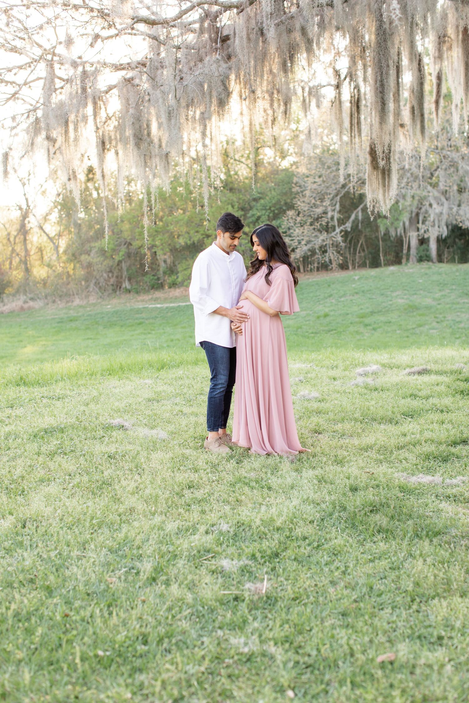 Couple shares intimate moment on grassy hill with Spanish moss draped trees in background.