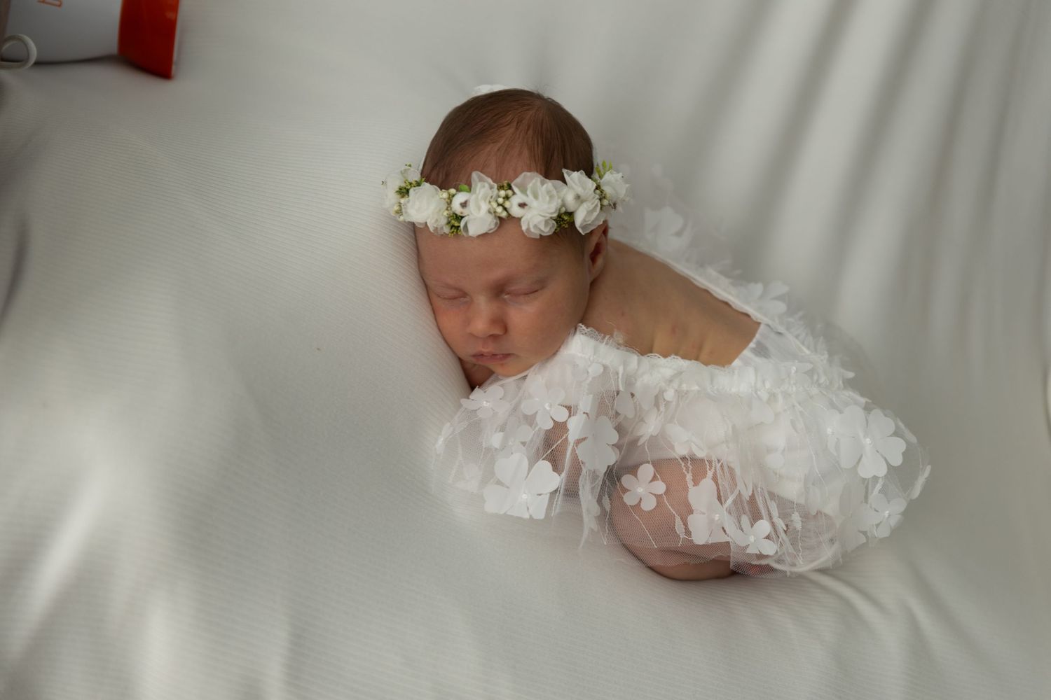 Sleeping newborn wearing white lace wrap and floral crown on soft white backdrop.