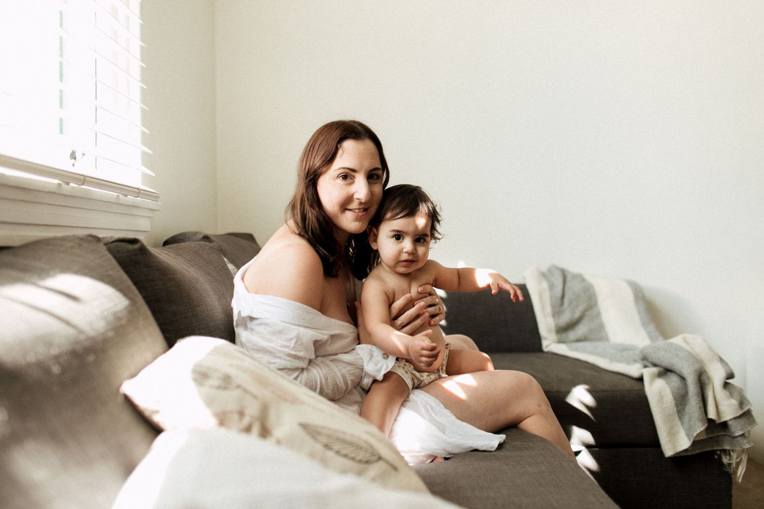 Young mother holding her baby on a grey couch in warm natural window light at home.