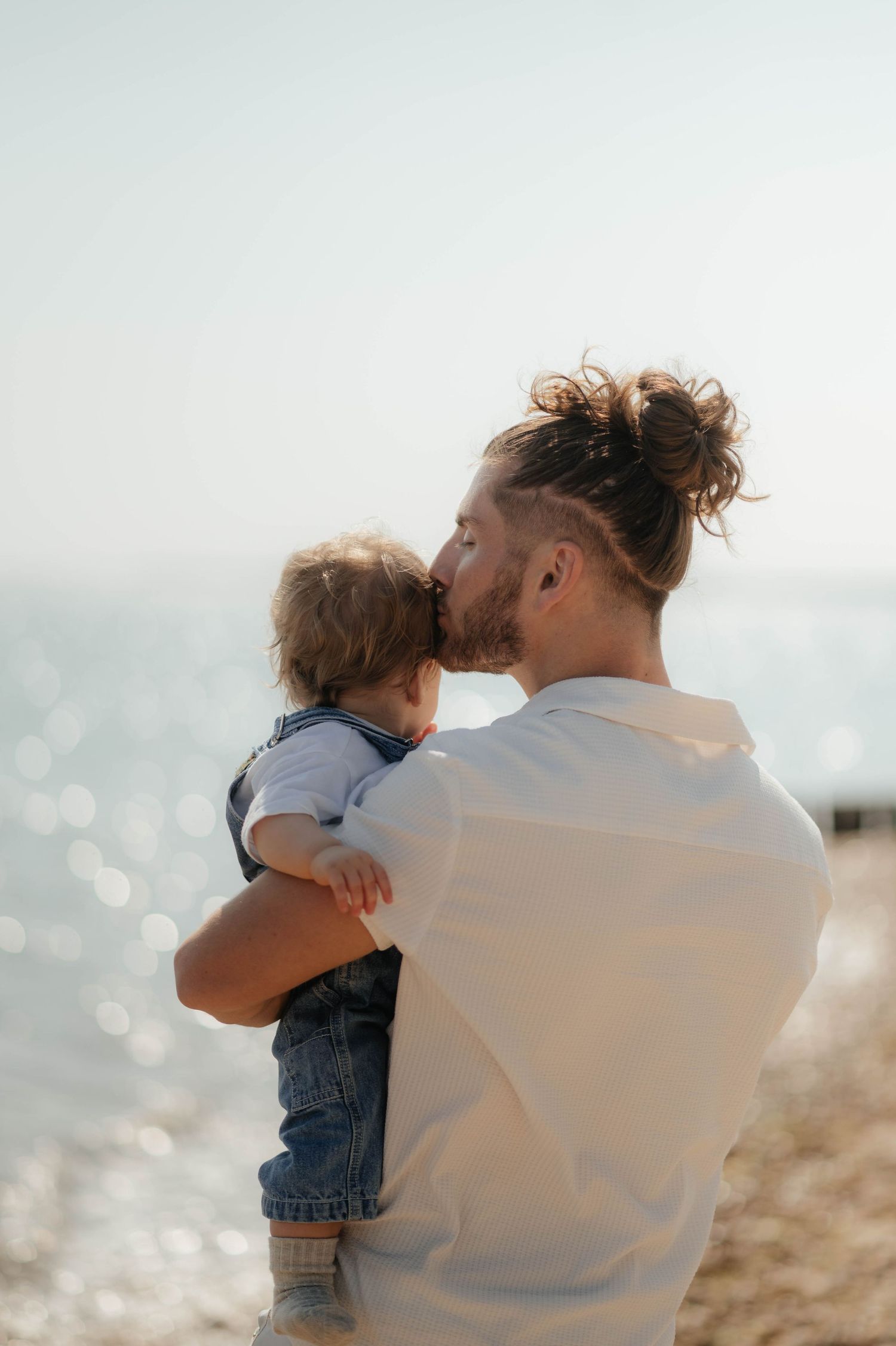 Parent in white shirt holding toddler by ocean with sun sparkling on water.
