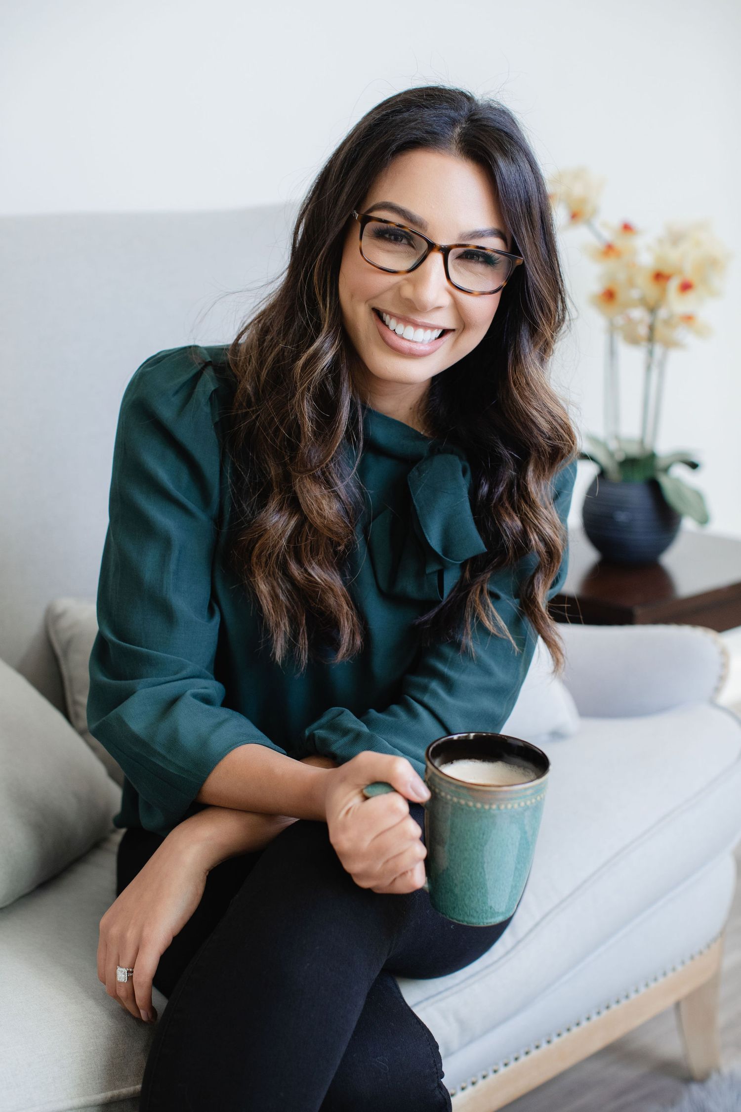 Smiling woman in green blouse wearing glasses holding coffee mug while sitting on couch.