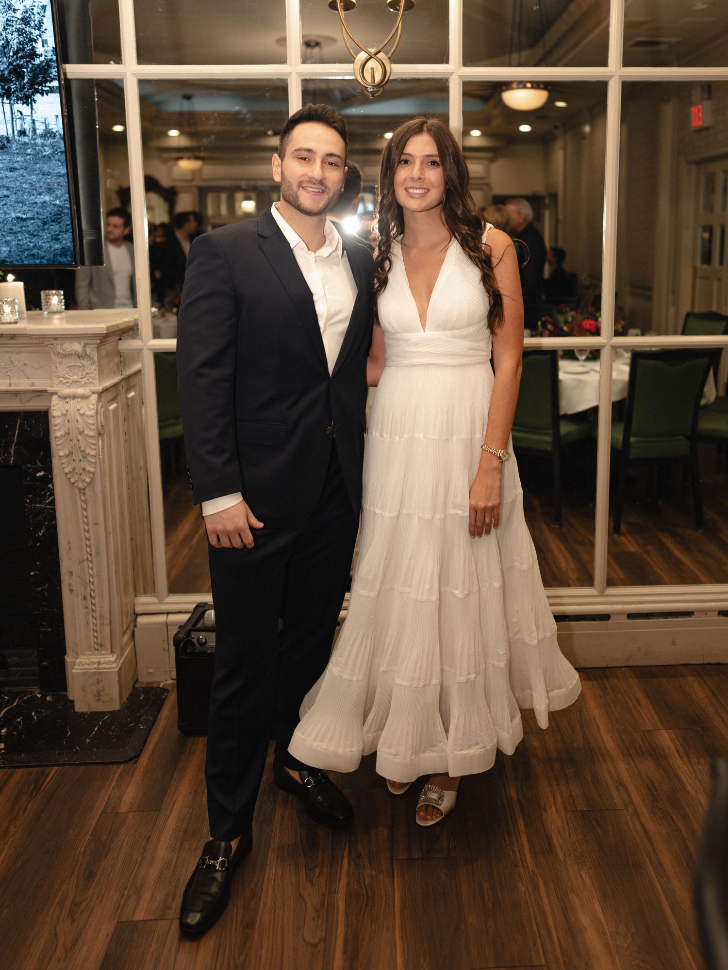 A formal couple posing against an elegant indoor backdrop with hardwood floors and windows.