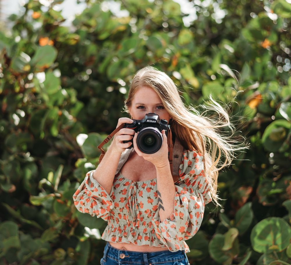 A photographer in a floral top captures images among fruit trees on a sunny day.