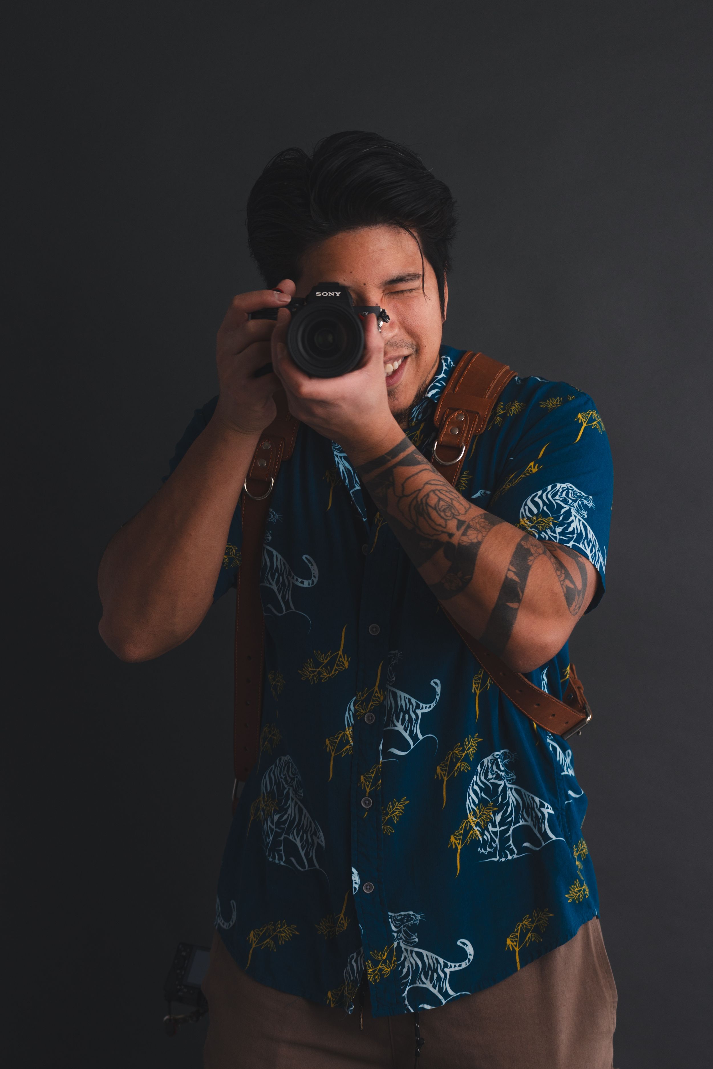 Photographer wearing blue patterned shirt takes photo with professional camera against dark background.