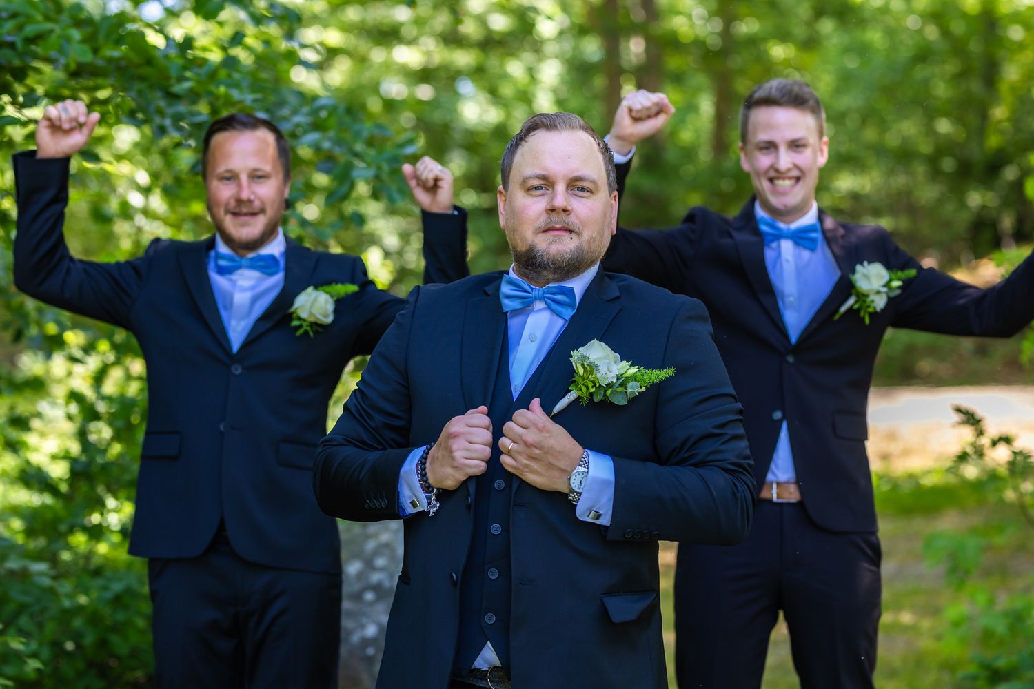 Groom and groomsmen in navy suits with blue bow ties celebrate outdoors, raising arms joyfully in a lush green forest setting.