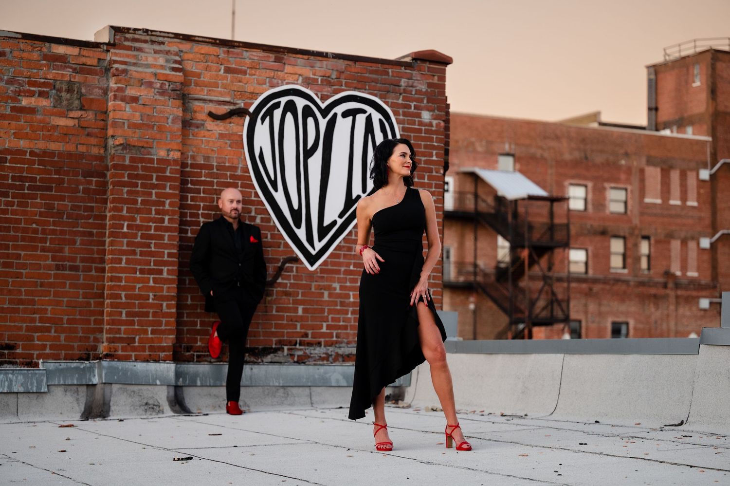 Two people pose dramatically against a brick wall with a heart graffiti design.