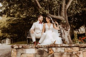 A romantic wedding couple sits together on a wooden bench in a garden setting.