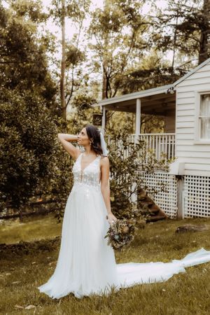 A flowing white wedding dress trails behind on the grass near a white cottage with lattice details.
