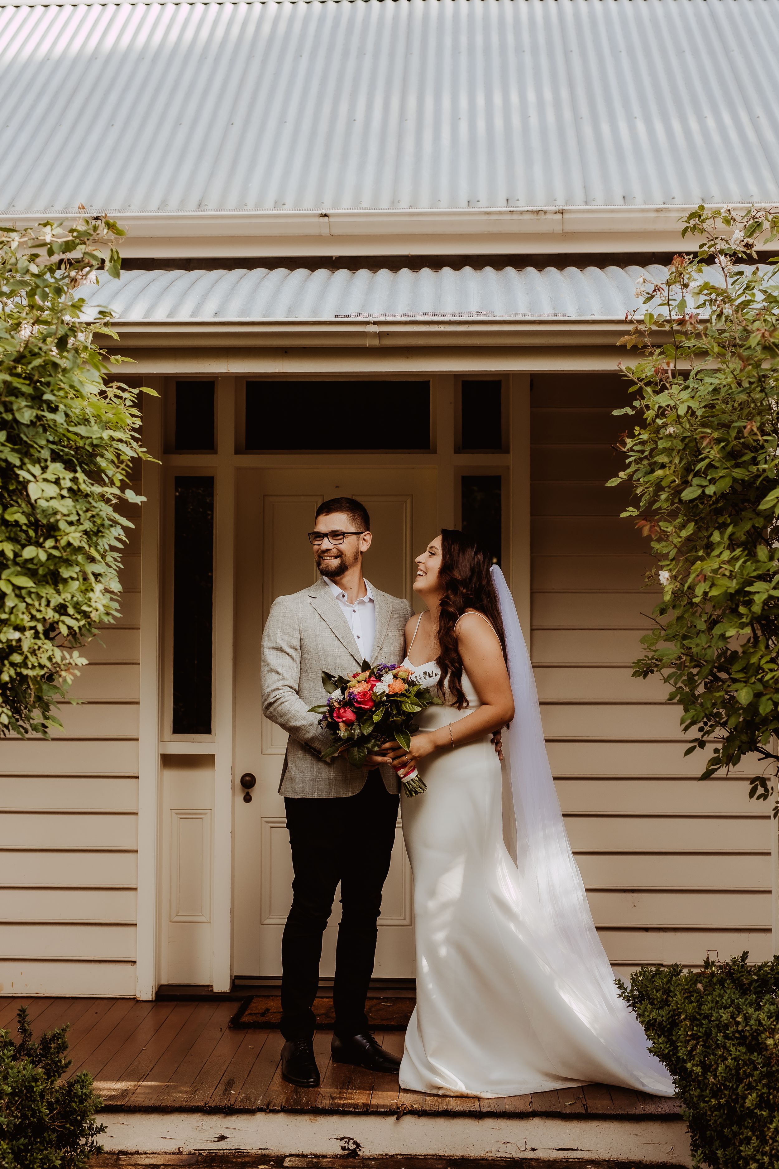 Newlyweds pose outside a white weatherboard house with garden bushes.