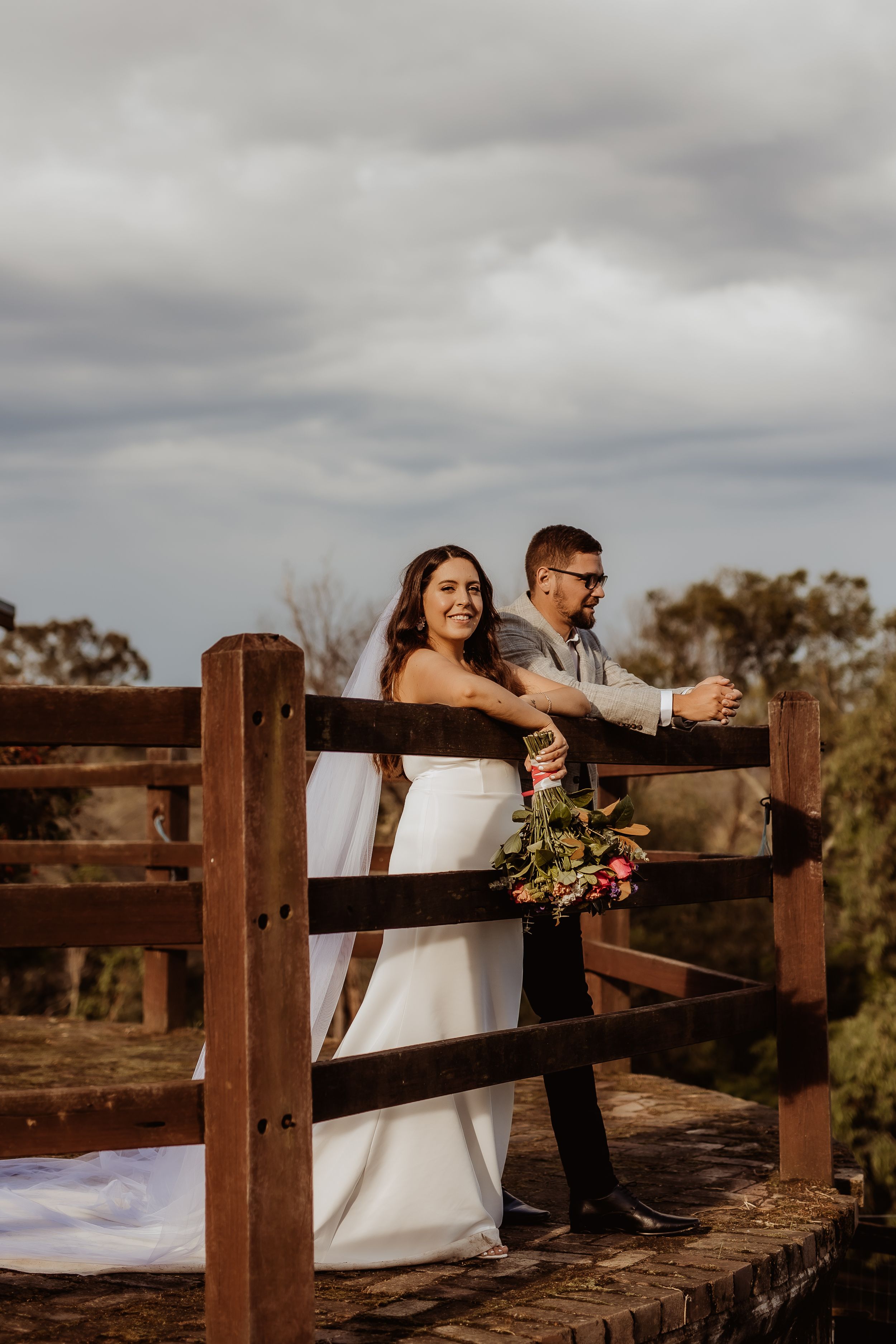 A romantic wedding photo series showing a couple leaning against a rustic wooden fence under cloudy skies.