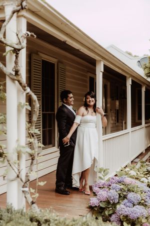 Wedding couple posing on white colonial porch with purple hydrangeas in foreground.