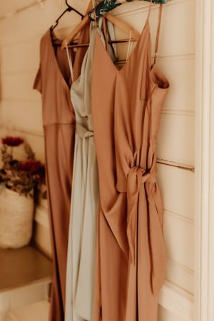 Pastel bridesmaid dresses in peach and mint tones hang elegantly on wooden hangers against a white wall.
