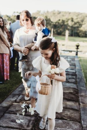 Young flower girls in white dresses carrying baskets during an outdoor ceremony.