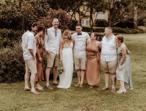 Wedding group photo with bridesmaids in dusty rose dresses and groomsmen in light beige suits.