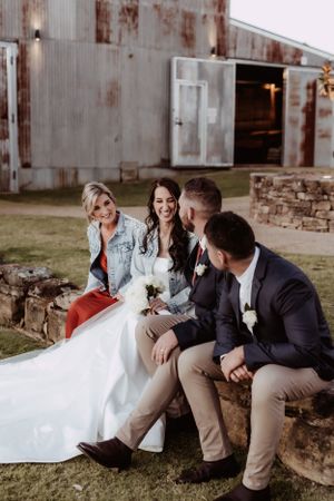 Wedding party sitting casually on stone wall sharing laughs during outdoor photo session.