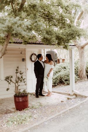 Elegant wedding couple sharing intimate moment on white columned porch surrounded by trees.