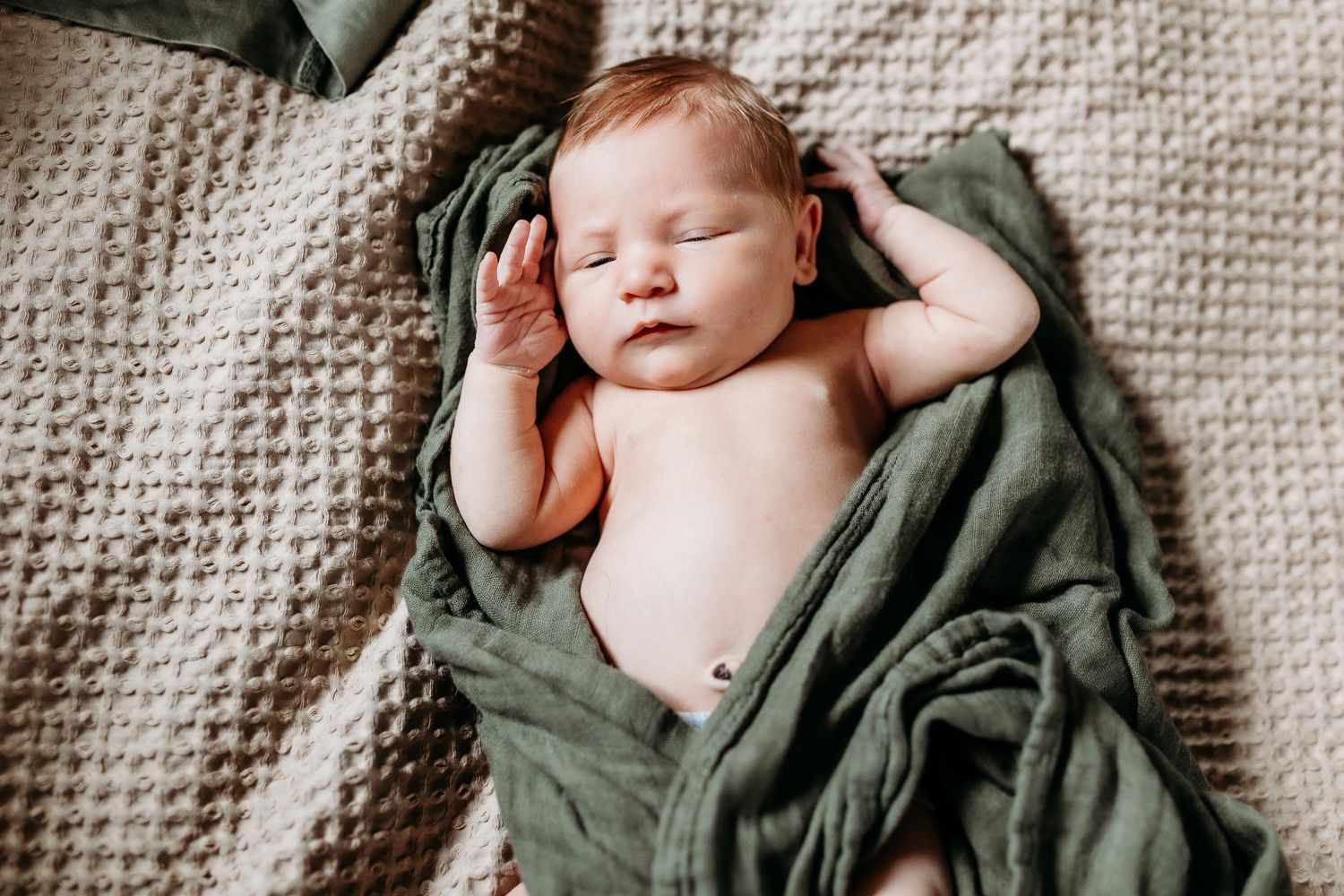 Sleeping newborn wrapped in olive green blanket on textured knit surface.