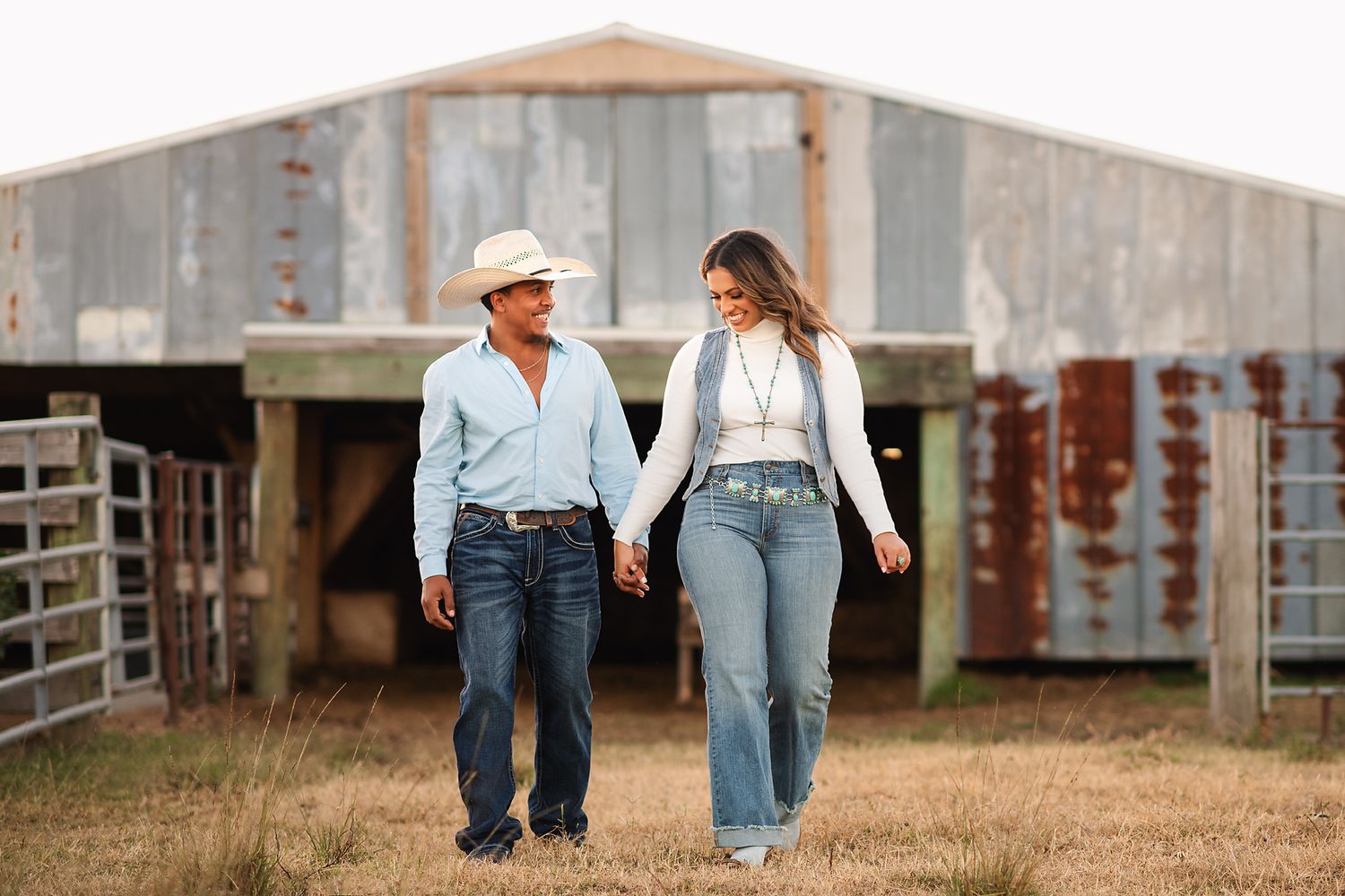 A couple walks hand in hand near a rustic barn wearing casual western attire.
